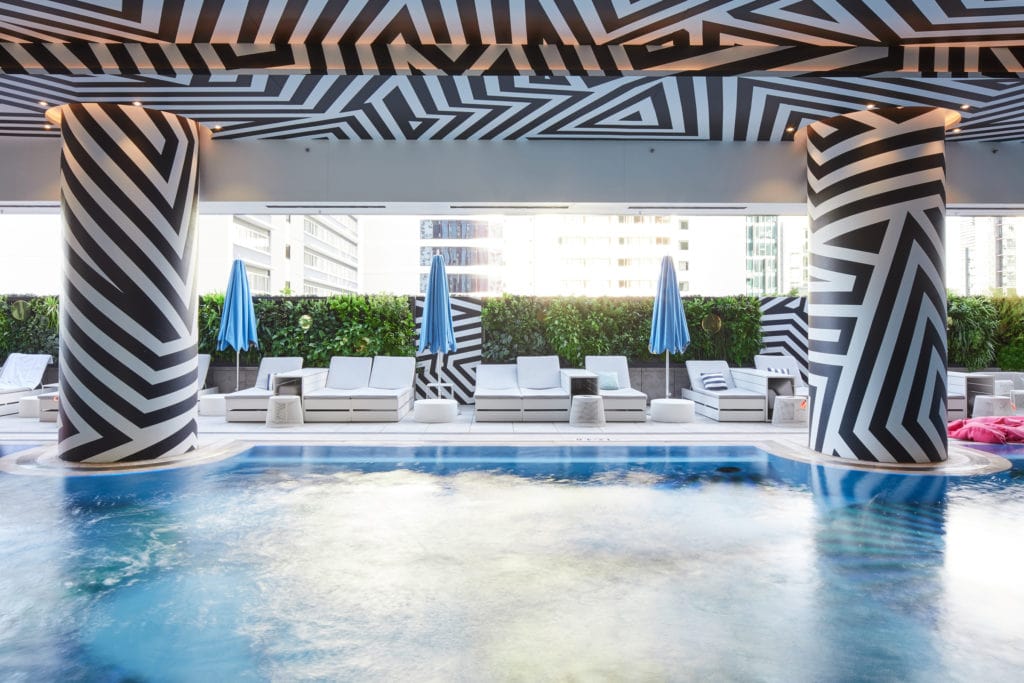 A green wall in our outside pool area at the W Hotel, Brisbane. In front of the green wall is three closed blue umbrellas, pool seating, and a well-maintained blue pool. Featuring striking black and white striped pillars and a ceiling.
