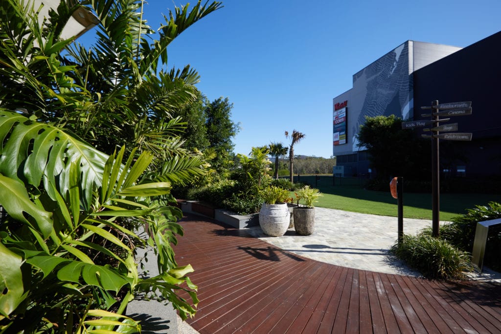 A professional photograph of a boardwalk in the outdoor area of retail shopping centre. The boardwalk has garden beds and potted plants all along it, with a Westfield sign in the background.