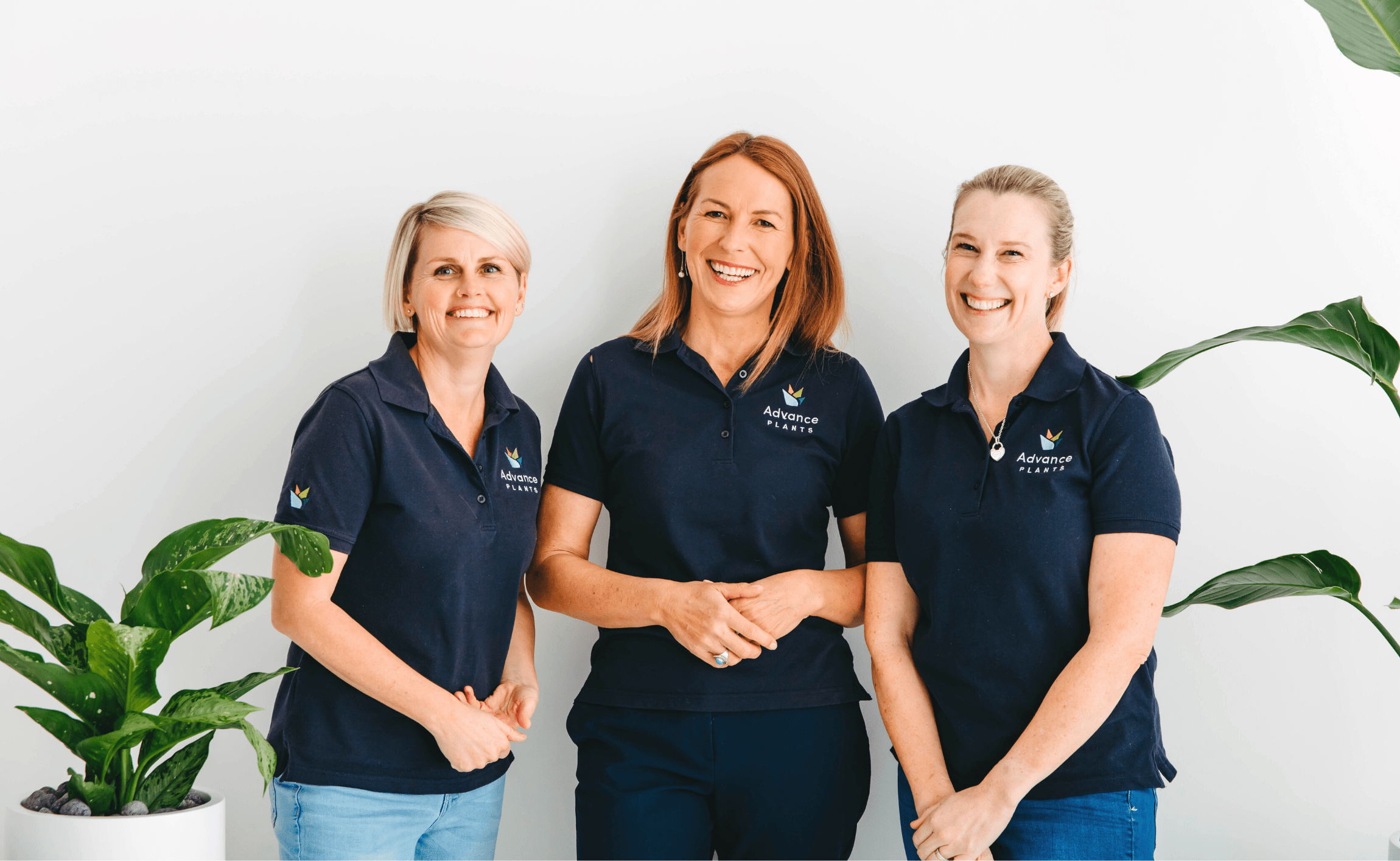 Kelli Forward, Sharon Prater and Amy Page of our indoor plant design team, bookended with potted plants. The ladies are smiling directly at the camera in their navy Advance Plants shirts.